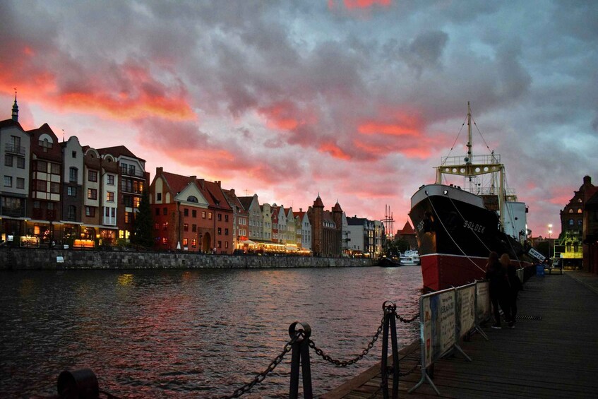 A waterfront cityscape with a sunset, a docked ship, and historic buildings. 1