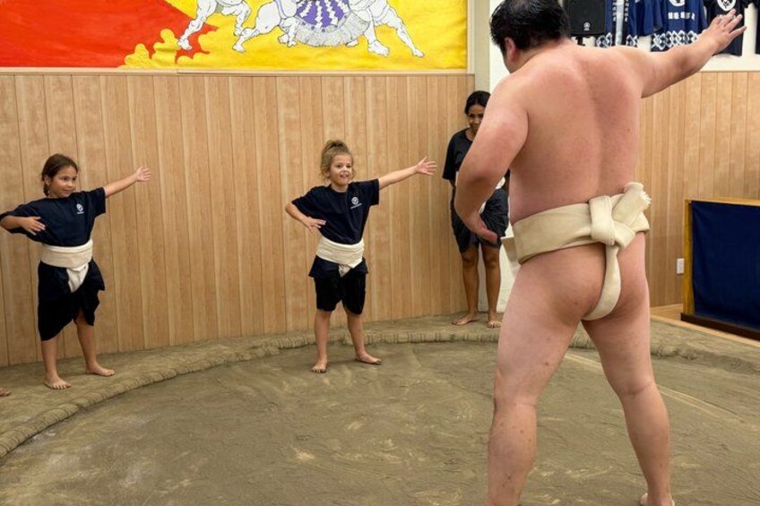 A sumo wrestler and two children in a training area with a wooden floor and a large sumo wrestling mural on the wall. 1