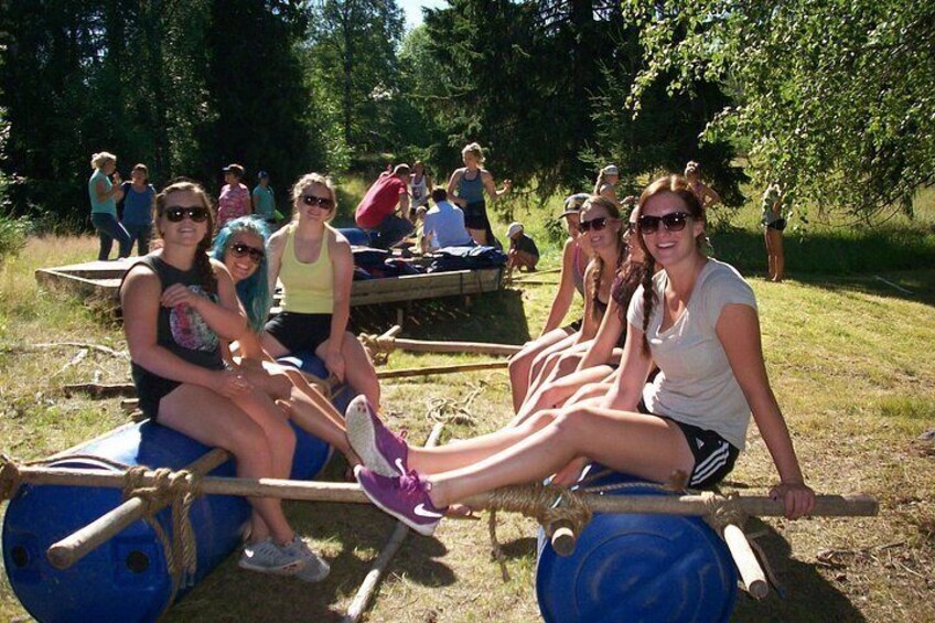 People sitting on a blue barrel with a rope across it, in a grassy outdoor area with trees. 1