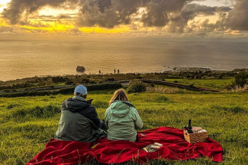 Two people sitting on a blanket overlooking a coastal landscape with a sunset. 2