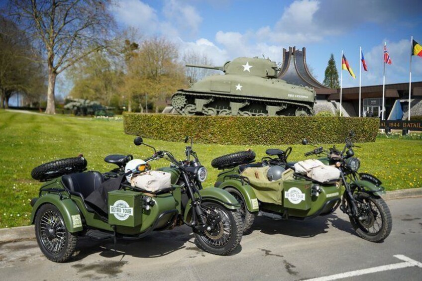 Two military motorcycles parked on a road with a tank and flags in the background. 4