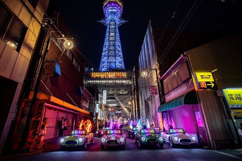 A brightly lit street with neon signs, a tower, and parked cars. 3