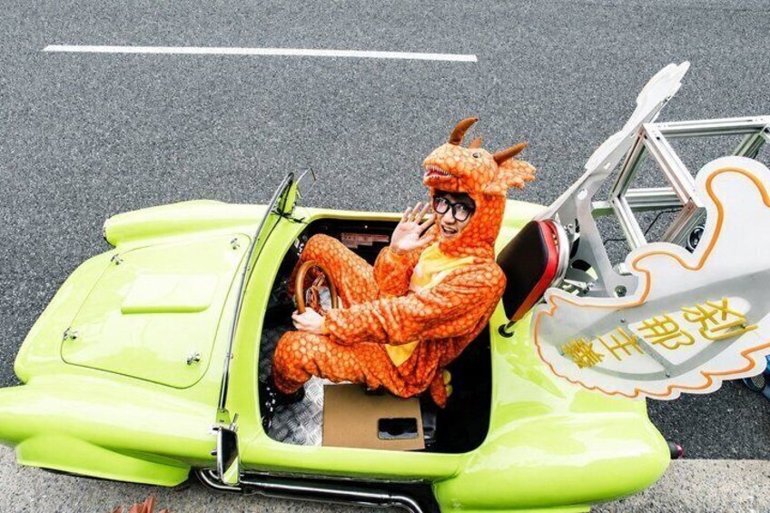 A person in a dinosaur costume sitting in a bright green car with a bicycle attached. 5