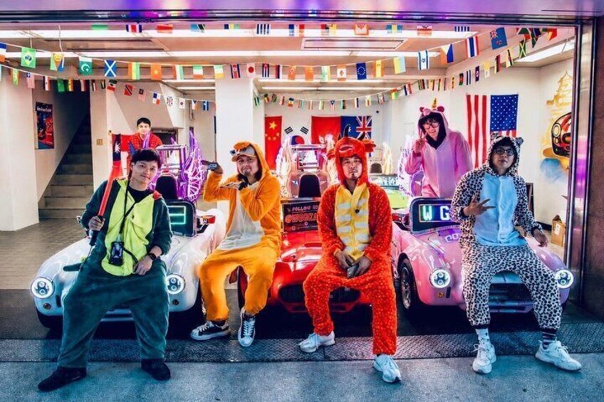 People in colorful costumes sitting on toy cars in a room decorated with flags. 3