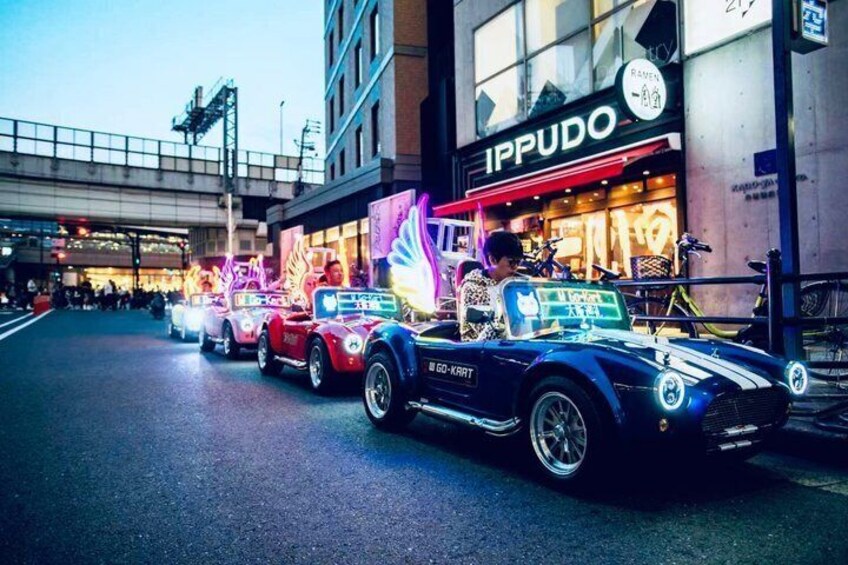 A row of classic cars parked on a street with illuminated signs and storefronts in the background. 1