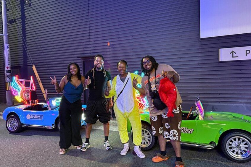 A group of people posing with a blue and a green car in front of a building with a sign. 5