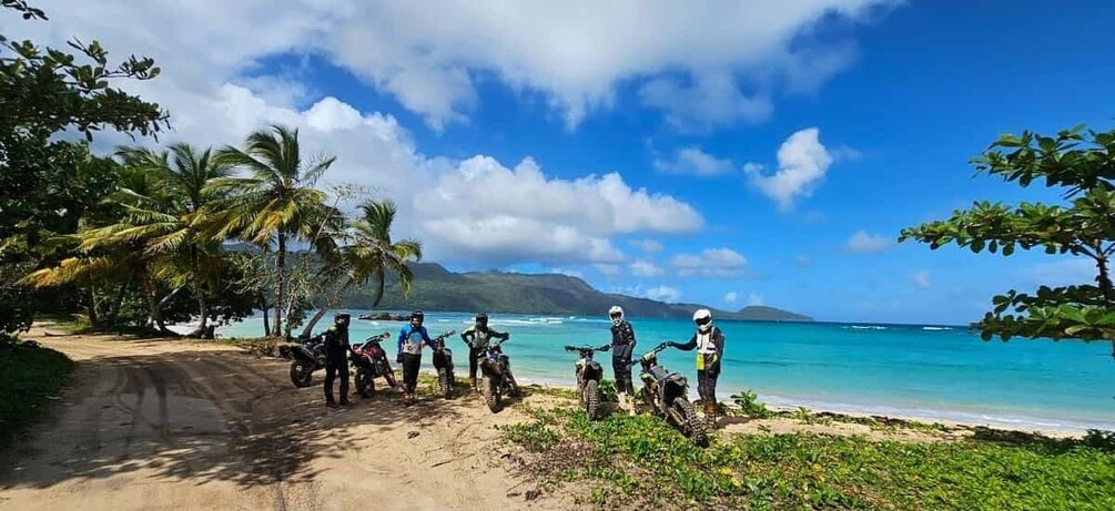 A group of motorcyclists on a dirt road by a beach with palm trees and a clear sky. 2