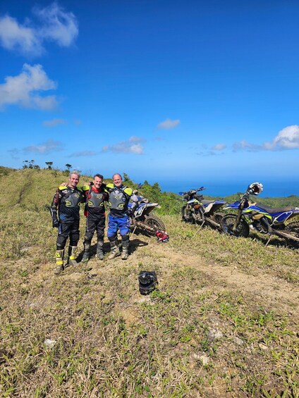 Two people in motocross gear standing on a grassy hill with a blue sky and ocean in the background. 3
