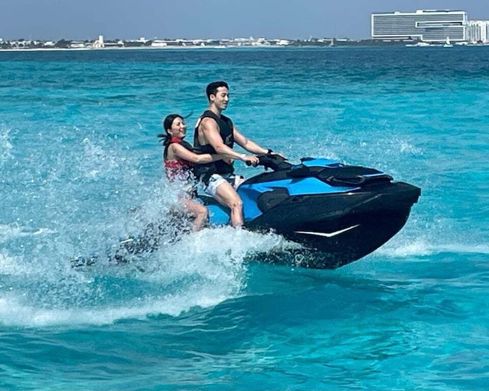 Two people jet-skiing on a clear blue sea with buildings in the background. 1