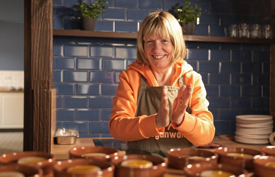 A person in an orange hoodie and apron standing in a kitchen with blue brick walls, surrounded by clay pots. 2