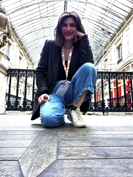 A person sitting on a wooden floor in a covered outdoor area with a glass ceiling, surrounded by ornate metal railings and classical architecture. 3