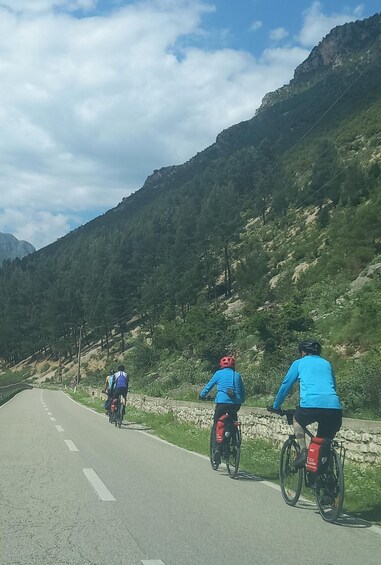 Three cyclists on a paved road with a mountainous backdrop. 1