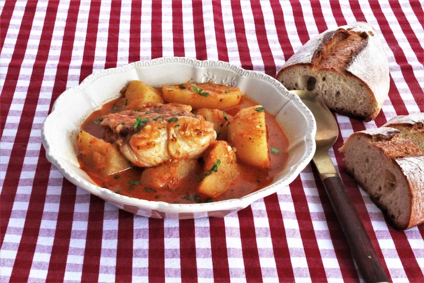 A bowl of stew with potatoes and meat, accompanied by slices of bread on a checkered tablecloth. 1