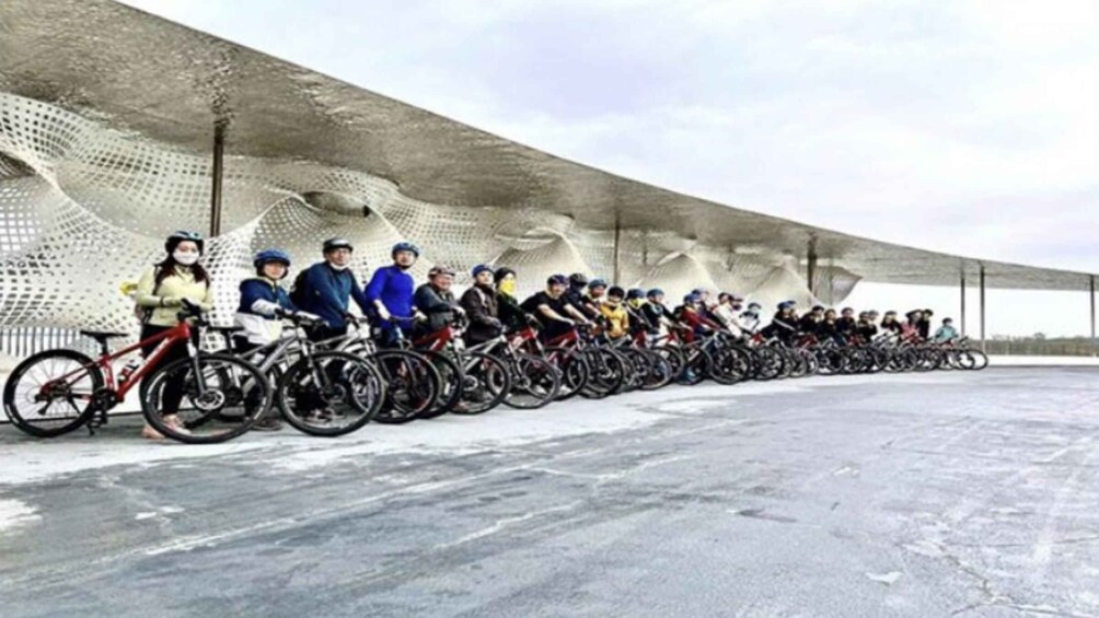 A group of cyclists lined up in a row on a concrete surface with a unique, wave-like architectural structure in the background. 4