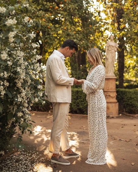 A couple holding hands in a garden with flowering bushes and a statue in the background. 2