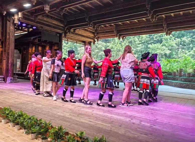 Eine Gruppe von Menschen führt einen traditionellen Tanz auf einer Bühne mit einer Holzkonstruktion und einem Hintergrund aus Bäumen auf. 5