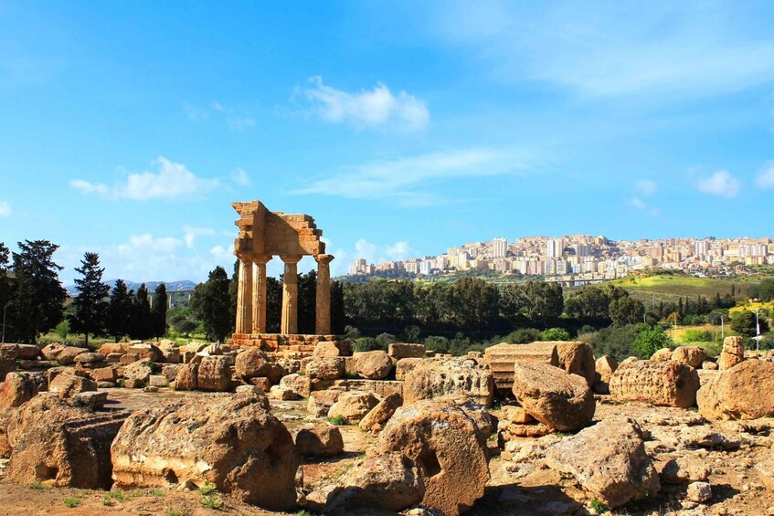 Ancient ruins with columns, large boulders, and a cityscape in the background. 3