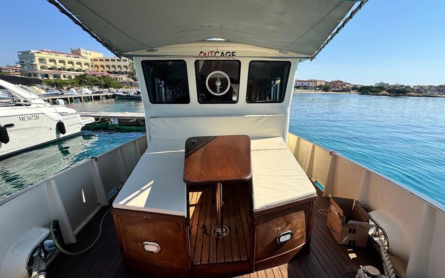 A boat's deck with a cushioned seating area, a steering wheel, and a view of the marina and buildings in the background. 2