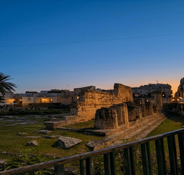 Ancient ruins with stone columns and steps, surrounded by modern buildings and a clear sky at dusk. 5