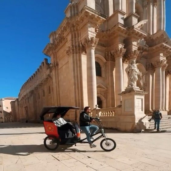 A historic building with ornate columns and statues, a person riding a bicycle, and another person sitting on a rickshaw. 4