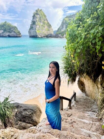 A person standing on a rocky path by a beach with clear turquoise water and green cliffs in the background. 5