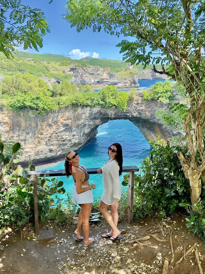 Two people standing by a cliff overlooking a blue sea, with greenery and a rocky landscape in the background. 2