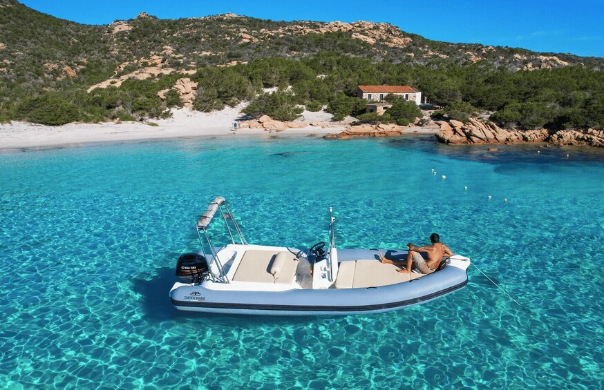 A clear blue sea with a boat anchored near a rocky shore, a white building on the hill, and a person sitting on the boat. 4