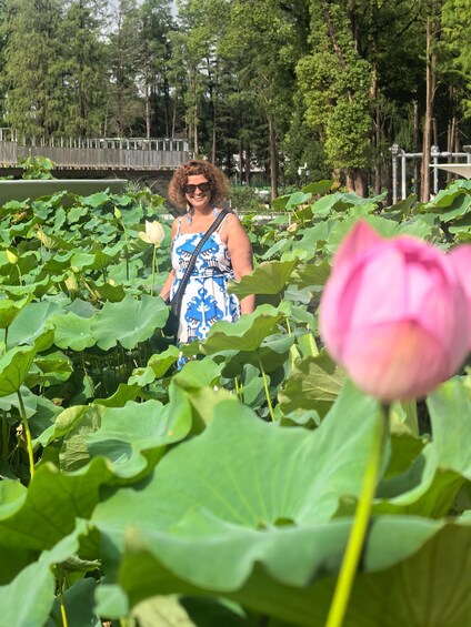 A person in a blue and white patterned dress walking through a lotus pond with large green leaves and a prominent pink lotus flower in the foreground. 1