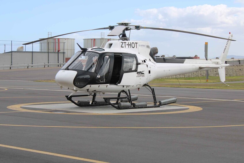 A white helicopter with 'ZT-HOT' and 'LICPOTERS' markings on a tarmac with a large industrial building in the background. 4