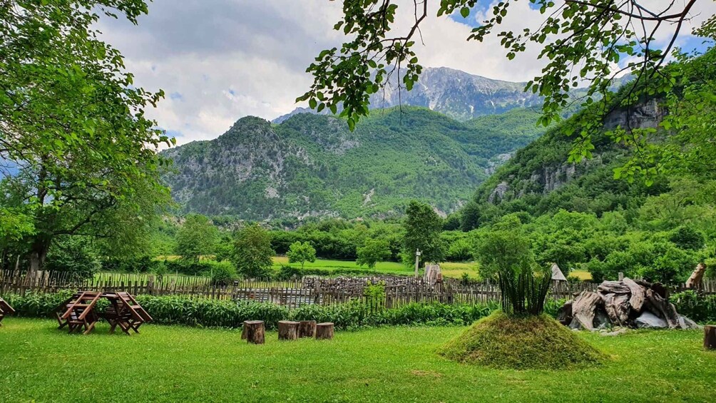 A lush green meadow with wooden chairs and a small tree stump, surrounded by a wooden fence and mountains in the background. 3