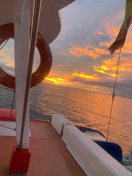 A boat deck with a lifebuoy and a flag, overlooking a sunset over the ocean. 4