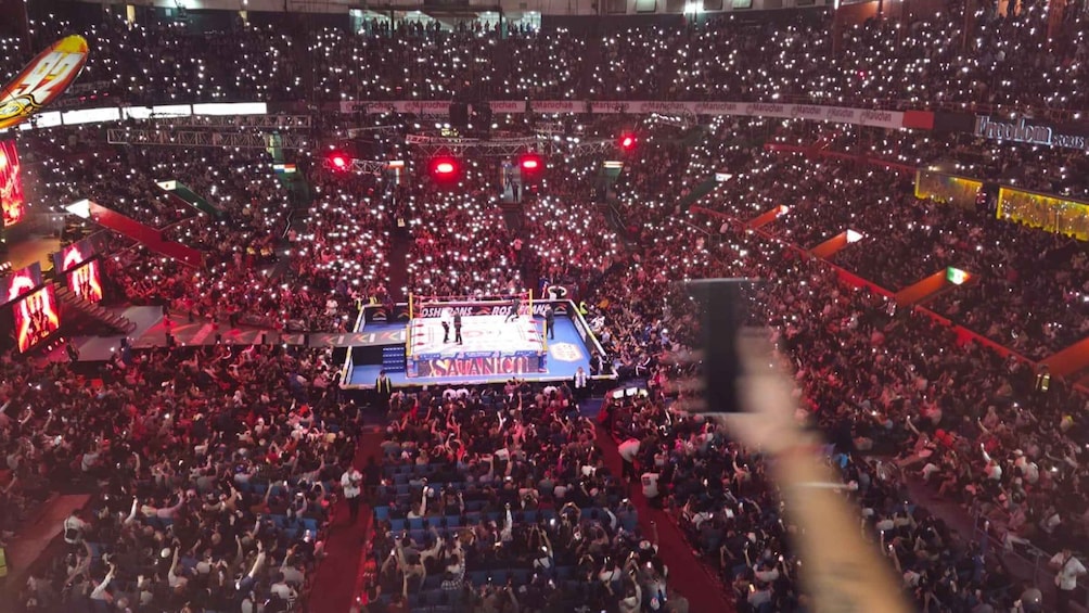 A crowded indoor arena with a stage at the center, surrounded by a sea of spectators holding up their phones. 3