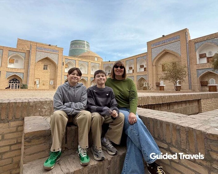 Three people sitting on a stone bench in front of a historic building with arches and domes. 2