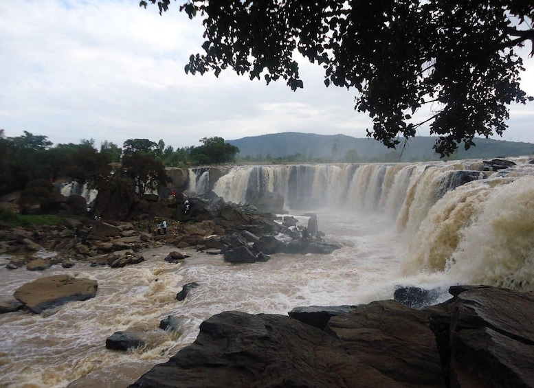 A multi-tiered waterfall with a rocky foreground and a forested backdrop. 2