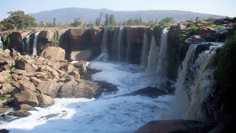 A multi-tiered waterfall cascading over rocky terrain with a river below, surrounded by hills and sparse vegetation. 4
