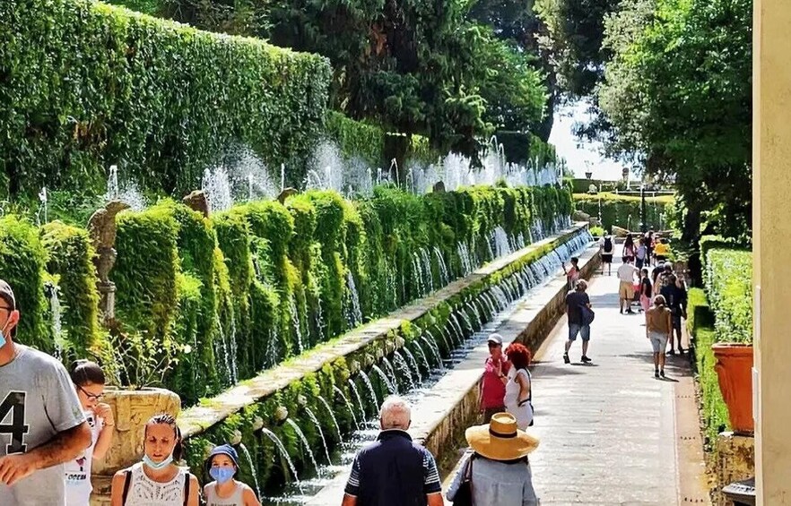 A garden with a long water feature, neatly trimmed hedges, and people walking along a path. 2