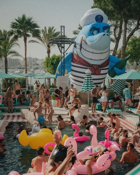 A crowded outdoor swimming pool with inflatable flamingos and a large inflatable character. 4