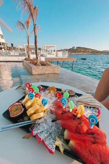 A table with a fruit platter, ice, and a view of a coastal area with palm trees and a white fence. 2