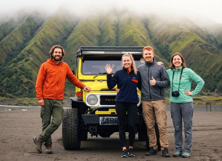 Four people standing in front of a yellow off-road vehicle with a mountainous landscape in the background. 2