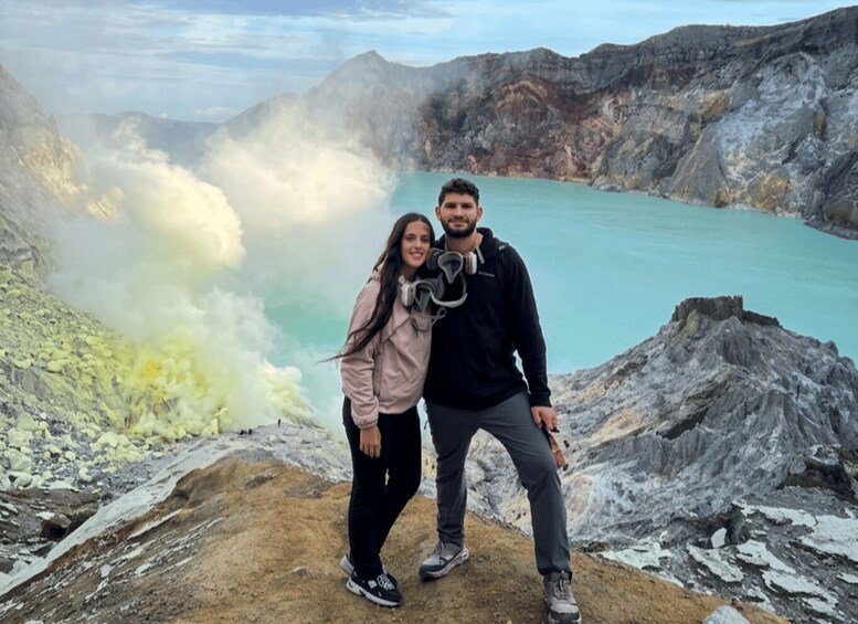 A couple standing on a rocky outcrop overlooking a turquoise lake with steam rising from the water. 4