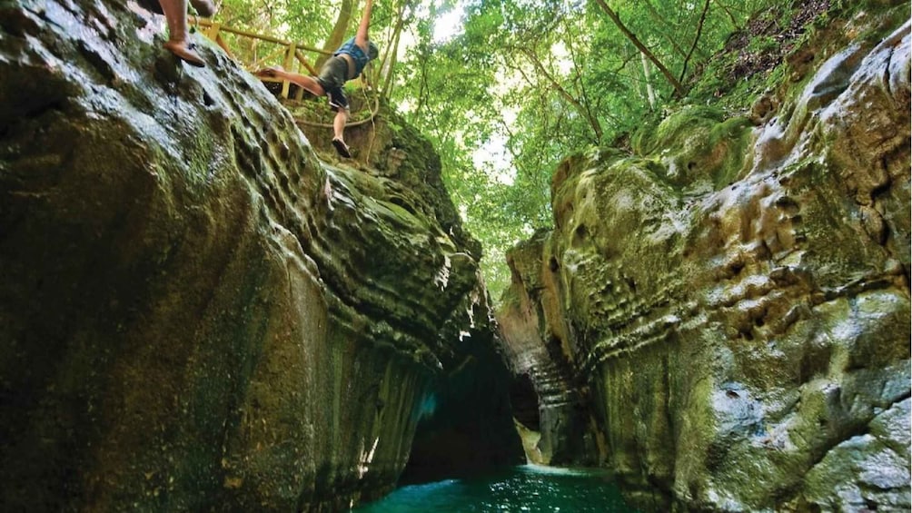 A person climbing a rocky cliff in a forested area with a narrow water channel below. 1