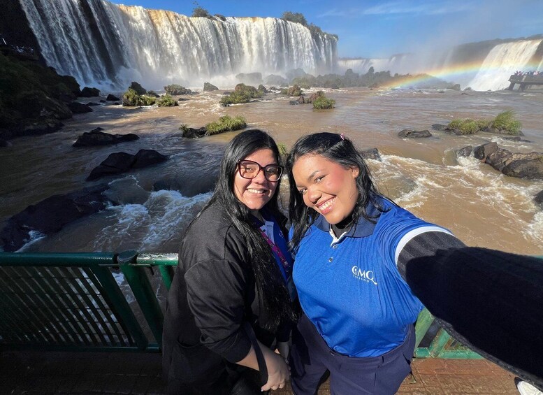 A waterfall with a rainbow, two people taking a selfie, and a river with rocks. 3