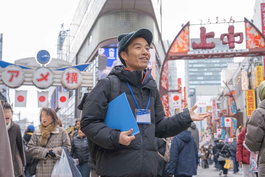 A person in a black jacket holding a blue folder, standing on a busy street with Japanese signage and buildings. 1