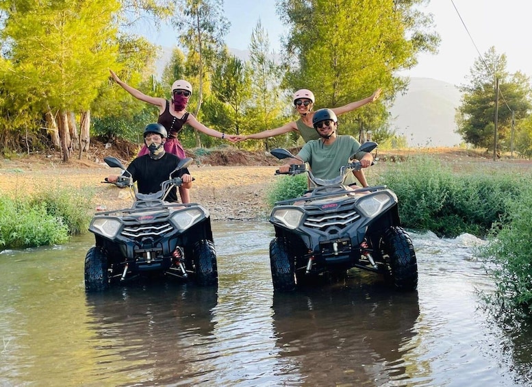 Two ATVs crossing a shallow stream in a wooded area. 1