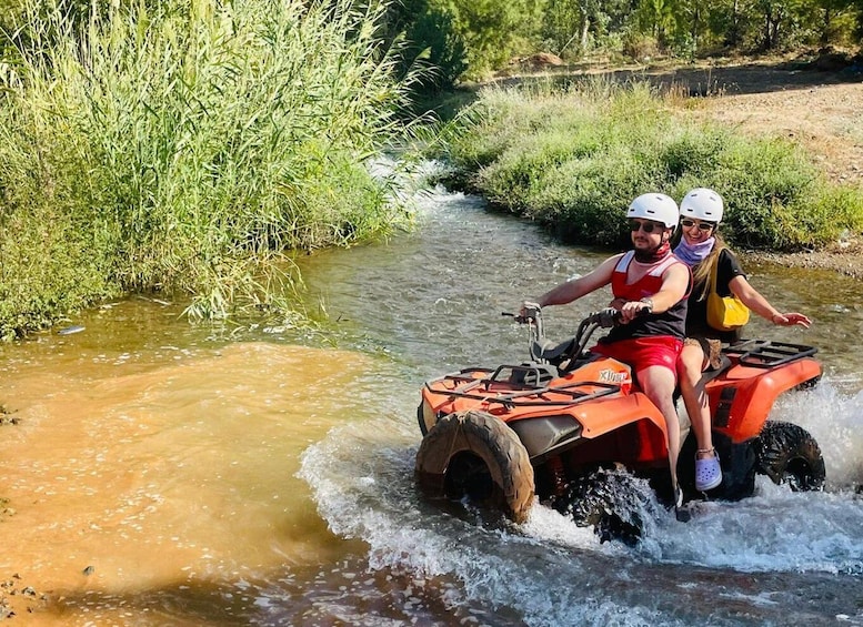 Two people on an ATV navigating a shallow stream surrounded by greenery. 2