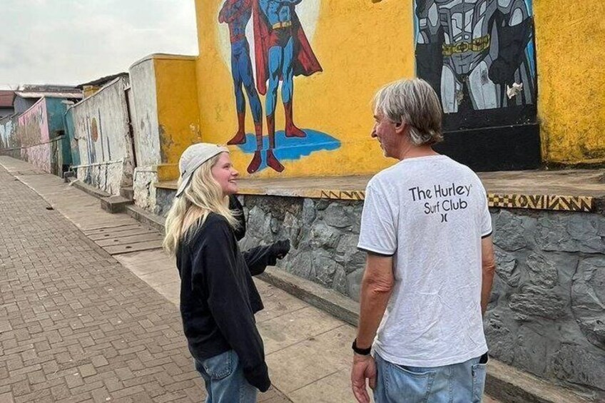 A person wearing a 'The Hurley Surf Club' t-shirt standing next to a mural of a superhero. 1