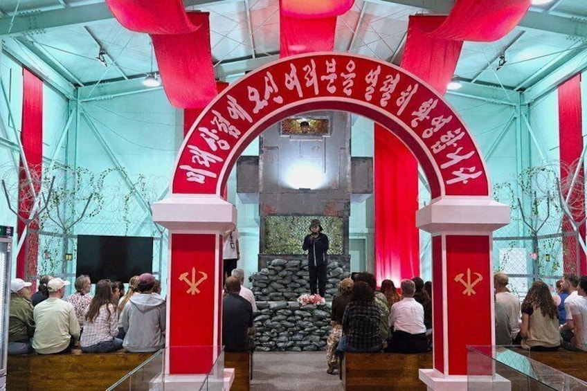 A group of people seated in a room with a red archway and a person standing at the front. 1