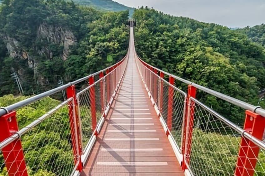 A suspension bridge with red railings, surrounded by lush green forest and rocky cliffs. 2