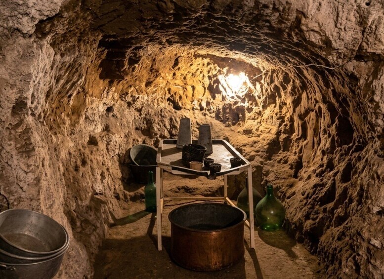 A dimly lit underground chamber with rough stone walls, a wooden table with pots, a metal bowl, and a green bottle. 3