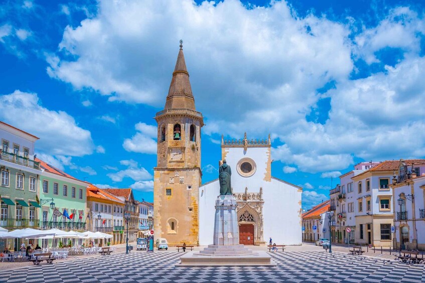 A historic square with a prominent clock tower, a white church, and surrounding colorful buildings. 2